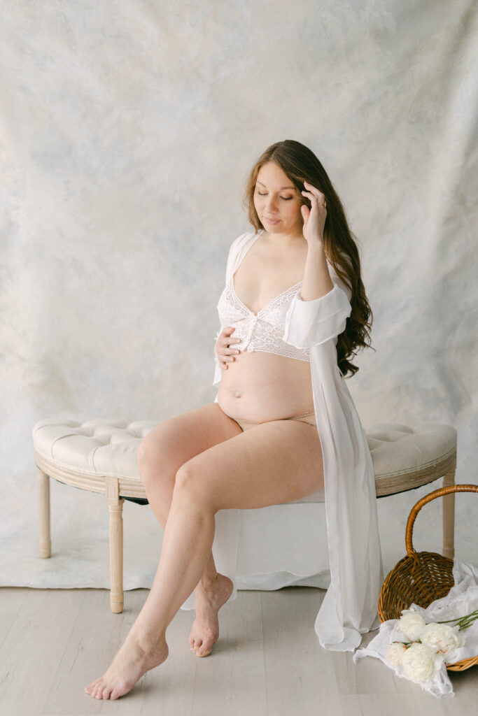 pregnant women sitting bench in front of fine art painted backdrop with one hand on swollen belly and the other in her hair. Trying to Get Pregnant in Knoxville TN. Knoxville maternity photographer