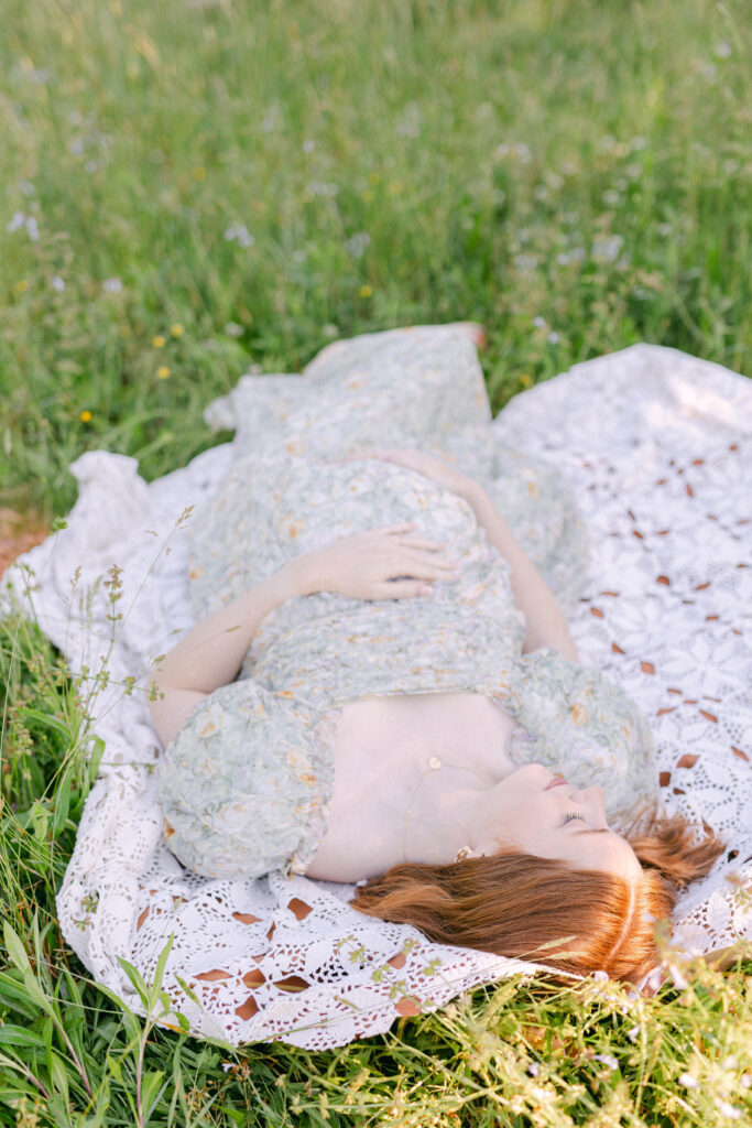 pregnant mom laying on knitted blanket in green pasture holding baby belly bump. Knoxville TN lifestyle fertility photographer